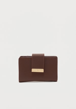 Cartera de cuero marrón con textura, con correa plegable y placa metálica dorada grabada con "ANNA FIELD" sobre un fondo blanco liso.