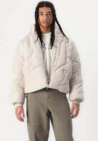 Veste matelassée beige avec capuche, dotée d'une fermeture éclair, de coutures texturées et de poignets élastiques, associée à un pantalon vert olive.