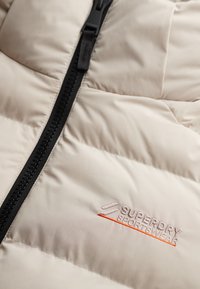 Chaqueta acolchada de color beige claro con cremallera negra. Presenta secciones cosidas y el logotipo bordado "Superdry Sportswear" en rosa y naranja.