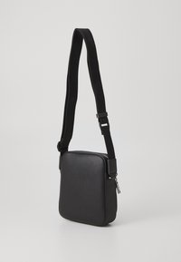 Borsa a tracolla nera realizzata in ecopelle testurizzata, caratterizzata da una forma quadrata, una tracolla in tessuto regolabile e dettagli in metallo argentato.