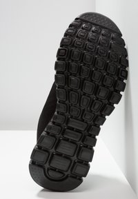 Semelle de basket noire avec des crampons texturés rectangulaires et carrés, présentée en gros plan sous un angle sur une surface blanche avec un arrière-plan blanc.