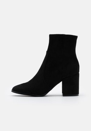 Bottine en daim noir avec un bout pointu et un talon bloc. Présente un design sans coutures avec des panneaux élastiques latéraux pour faciliter l'enfilage.