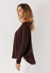 Maglia marrone a maniche lunghe con taglio rilassato, tessuto morbido e orlo arrotondato. Indossata sopra pantaloni bianchi, con una leggera caduta e spacchi laterali.
