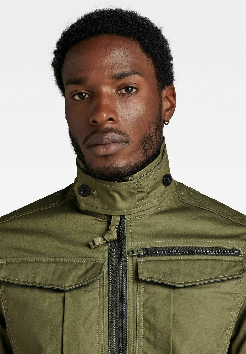 Giacca verde oliva con colletto alto, zip frontale, due tasche sul petto e cuciture decorative. Presenta una trama di tessuto robusto e accessori neri.