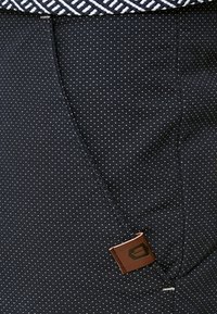 Tissu sombre à petits pois blancs, coutures visibles et étiquette en cuir marron près de la couture sous une ceinture élastique rayée.