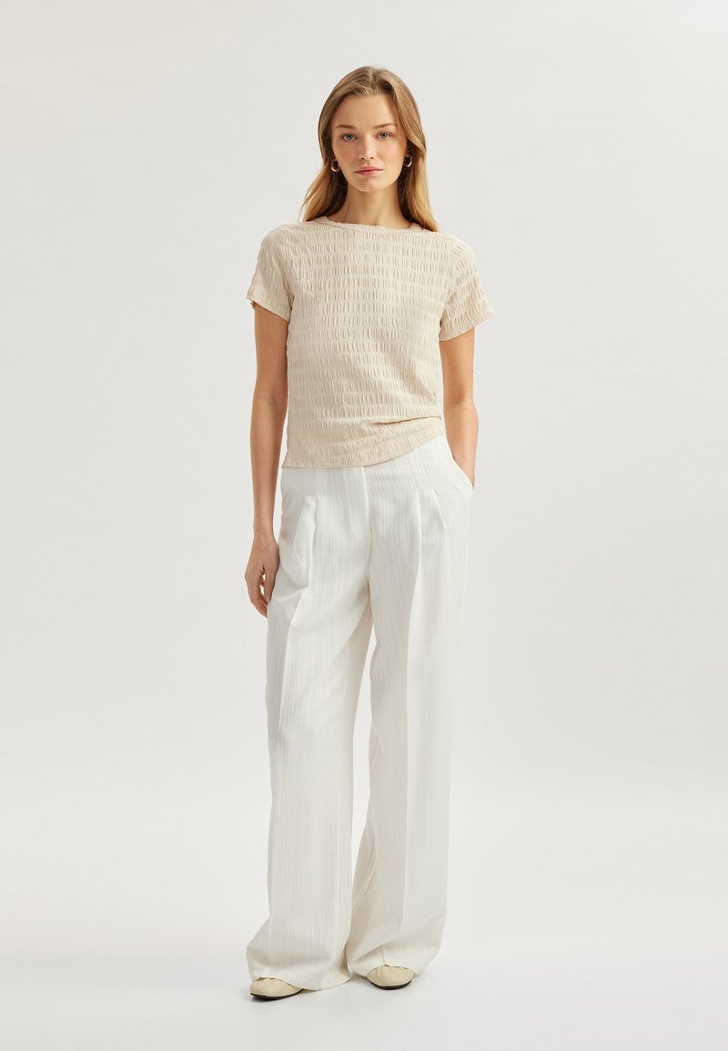 Top beige chiaro testurizzato con maniche corte abbinato a pantaloni bianchi a gamba larga plissettati. Include un tessuto morbido e liscio con dettagli sottili.