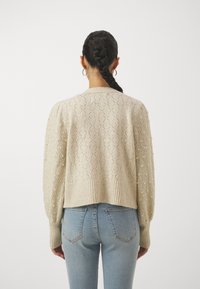 Beżowy sweter z dzianiny o fakturowanym wzorze, z prążkowanym dołem i bufiastymi rękawami. Tył swetra ma lekko skróconą długość.