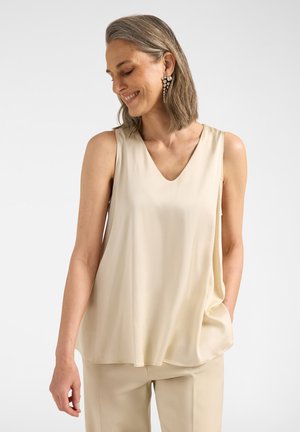 Vrouw met grijs haar, pareloorbellen, een mouwloos beige topje en bijpassende beige broek, glimlachend en naar beneden kijkend.