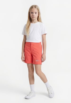 Mladé dievča stojí, obliekané v bielom tričku, korálových šortkách s logom, bielych ponožkách a bielych teniskách na jednofarebnom bielom pozadí.