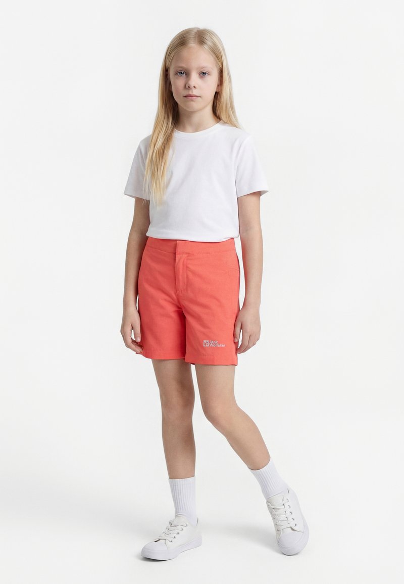 Νεαρή κοπέλα στέκεται, φοράει άσπρο μπλουζάκι, κοραλλί σορτς με λογότυπο, λευκές κάλτσες και λευκά αθλητικά παπούτσια σε απλό λευκό φόντο.