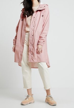 Woman wearing a pink raincoat over a light sweater, cream cropped pants, and beige sneakers standing against a plain background.