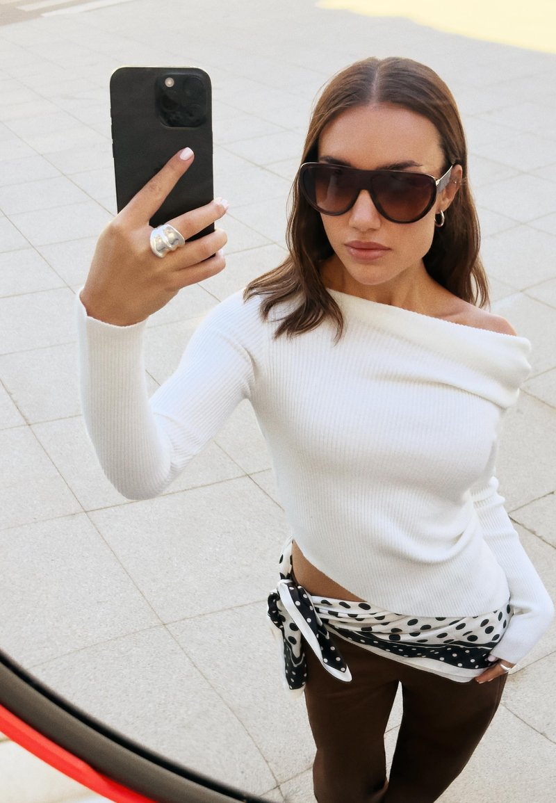 Bela rebrasta bluza z dolgimi rokavi in off-shoulder obliko, v kombinaciji s sivimi hlačami in črno-belim pikastim šalom. Velike sončna očala.