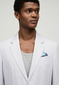 White blazer with notched lapels over a gray t-shirt, featuring a patterned pocket square with blue accents. Smooth, lightweight fabric.