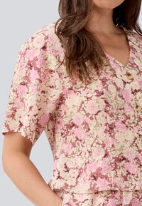 Blusa floreale con maniche corte, caratterizzata da un motivo rosa, crema e marrone. Design a V con tre bottoni in perla sul davanti. Tessuto testurizzato.