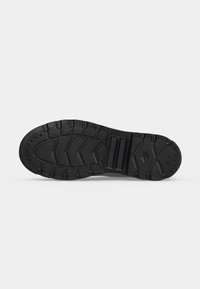 Semelle de chaussure en caoutchouc noir avec des motifs de bande de roulement profonds et des rainures rectangulaires et en forme de chevrons pour l’adhérence sur fond gris clair.