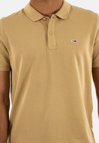 Polo de tela en tono beige con un acabado texturizado. Presenta un color clásico, dos botones y un pequeño logo en el lado izquierdo del pecho.