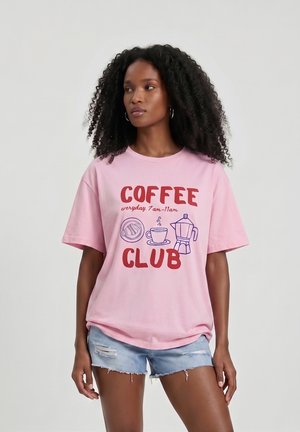 Kobiet krussis juustega, kellel on seljas roosa "Coffee Club" T-särk ja rebenenud teksapüksid, ees hele ühtlane taust.