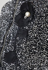 Giacca nera con paillettes e chiusura a zip, presenta un декоративità in tessuto a forma di fiore e perle, con dettagli in satin. Superficie lucida e testurizzata.