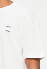 White cotton t-shirt with a crew neck. Features small, dark blue "SAMØE" text embroidered twice on the left side of the chest.