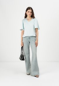 Top azzurro chiaro a V con maniche a sbuffo abbinato a jeans chiari a gamba svasata. La modella tiene in mano una piccola borsa nera, in piedi contro uno sfondo bianco.