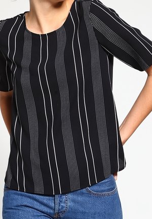 Nainen, jolla on päällään musta lyhythihainen pusero, jossa on pystysuoria valkoisia pinstripes-raidat, ja siniset farkut, seisoo yhdellä kädellä lantiolla.