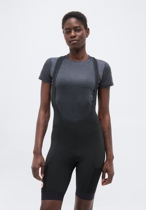 Tânără femeie de culoare purtând un tricou gri mulat și pantaloni scurți de ciclism negri cu bretele, stând pe un fundal simplu și deschis la culoare.