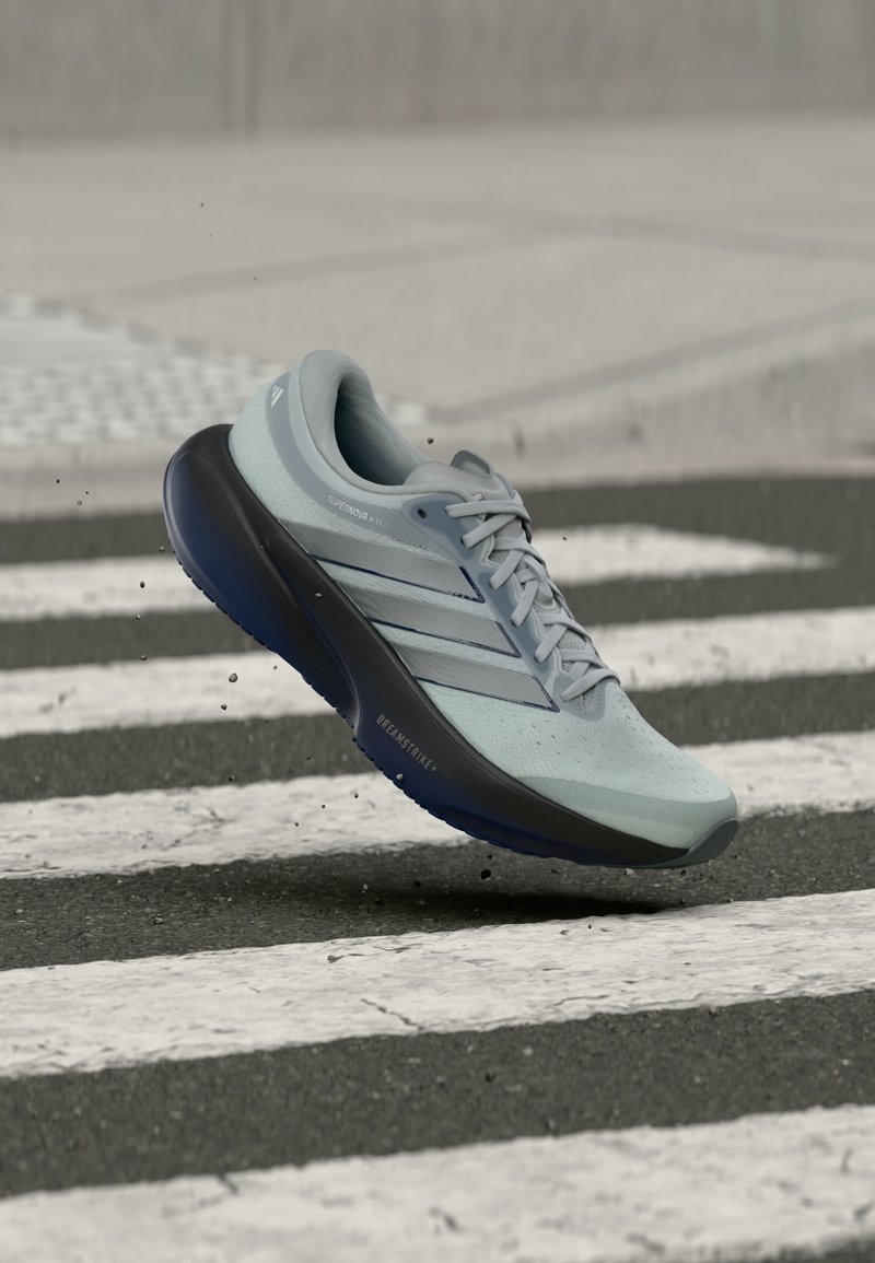 Ανοιχτό μπλε παπούτσι τρεξίματος Adidas με μαύρη σόλα, στον αέρα πάνω από διάβαση πεζών σε άσφαλτο, εμφανίζοντας τις ρίγες της μάρκας και λεπτομέρειες κορδονιών.