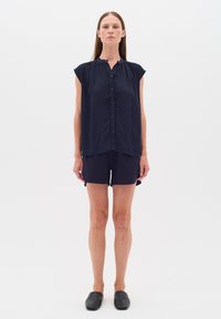 Marinblå ärmlös blus med knappfront och rynkad halsringning, kombinerad med shorts. Slät tyg, enkel design, minimal detaljer.