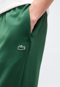 Άτομο που φοράει πράσινο σορτς με το χέρι στην τσέπη, το οποίο έχει ένα μικρό κεντημένο λογότυπο κροκόδειλου στο πλάι.