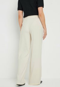 Pantalones de pierna ancha en tejido crema claro, con cintura alta y una textura suave. La vista trasera muestra costuras entalladas y un sutil drapeado.