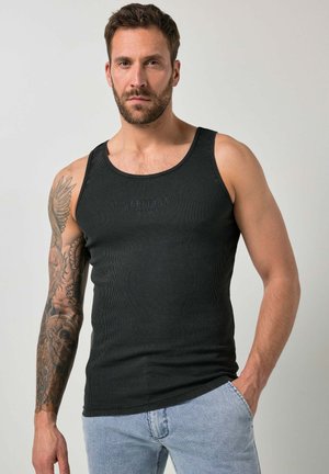 Mężczyzna z brodą i tatuażem na całym lewym ramieniu, noszący czarny tank top i jasnoniebieskie dżinsy, pozujący z prawą ręką w kieszeni.