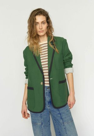 Femeie cu părul ondulat poartă un sacou verde cu tiv negru, o cămașă cu dungi și blugi albaștri, stând într-un cadru simplu, cu fundal deschis.