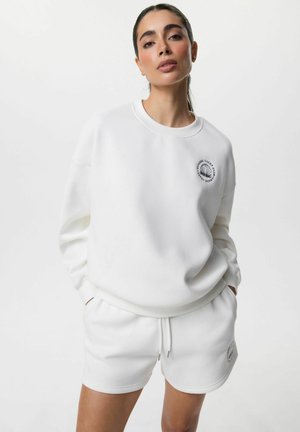 Kobieta o ciemnych włosach, ubrana w białą bluzę i dopasowane białe szorty z małymi czarnymi okrągłymi logo, stoi z rękami w kieszeniach.