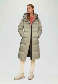 Lange, lichtgrijze pufferjas met capuchon, voorzien van drukknoopsluiting en zijzakken, gecombineerd met een roze trui en beige enkellaarsjes.