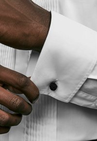 Camicia bianca con design a pieghe e gemello a nodo nero. Primo piano di una mano che sistema il polsino. Tessuto liscio e vestibilità su misura.