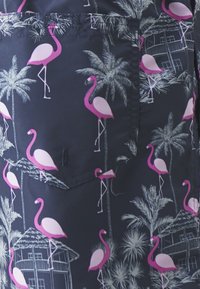 Tejido azul marino con un estampado tropical que incluye flamencos rosas y palmeras. Incluye un bolsillo con detalles cosidos y formas geométricas.
