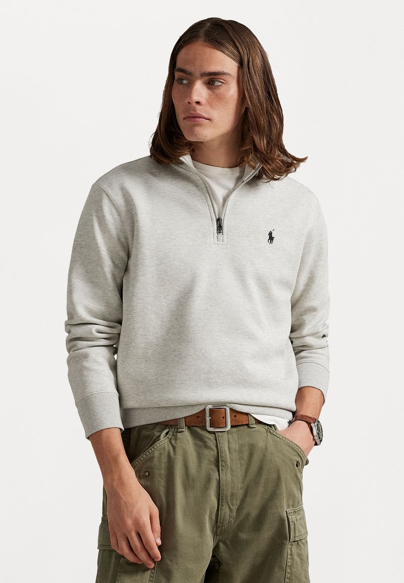 Ljusgrå zip-up sweatshirt med ribbade ärmar, i kombination med olivgröna cargobyxor och ett brunt bälte. Har en liten broderad logotyp på bröstet.