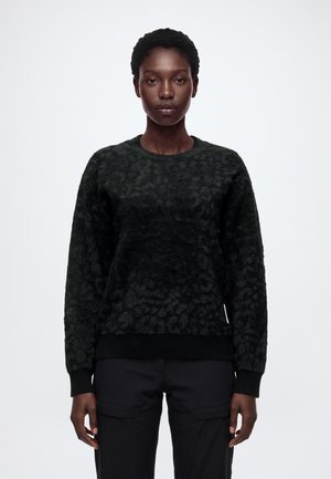 Un mannequin se tient devant un fond blanc uni, portant un pull foncé à motifs et un pantalon cargo noir.
