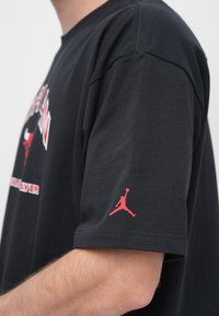 Μαύρο βαμβακερό μπλουζάκι με κοντά μανίκια, με κόκκινο κείμενο και λογότυπο Jumpman στο αριστερό μανίκι. Απλός, άνετος σχεδιασμός.