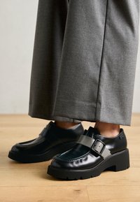 Chaussures en cuir noir avec une semelle épaisse et des accents de sangle gris. Forme arrondie avec des détails de couture et une finition brillante. Présentées avec un pantalon gris.