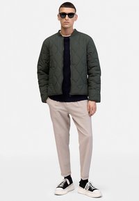 Quilted green bomber jacket with a round neck, worn over a navy sweater; paired with light trousers and black sneakers. Black sunglasses worn.