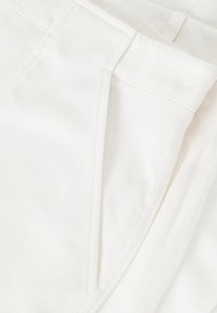 Tessuto bianco con una consistenza liscia, caratterizzato da cuciture sartoriali e un design pulito. Il capo include tasche e dettagli di cucitura precisi.
