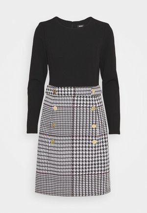 Haut noir à manches longues avec jupe pied-de-poule noir et blanc attachée, présentant une double rangée de boutons dorés à l'avant.