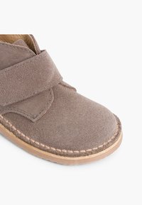 Scarpa in suede marrone con punta rotonda e cinturino regolabile in velcro, dotata di suola in gomma leggermente tan e dettagli di cucitura visibili.