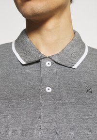 Siva polo majica s teksturiranim uzorkom, bijelim detaljima na ovratniku i tri bijele gumbe. Ima mali crni logo na prsima.