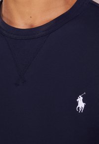 Närbild av en marinblå klädesplagg med synlig sömnad och en vit broderad polo-spelare logotyp på bröstet.