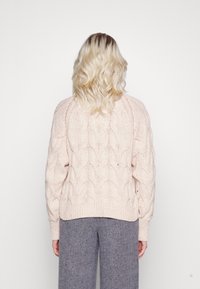 Personne blonde portant un pull en maille torsadée crème et un pantalon gris, se tenant de dos face à la caméra sur un fond uni.