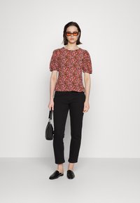 Blusa com padrão floral em tons de rosa, vermelho e amarelo; mangas curtas com franzido; bainha reta. Combinada com calças ajustadas pretas e sapatos pretos.