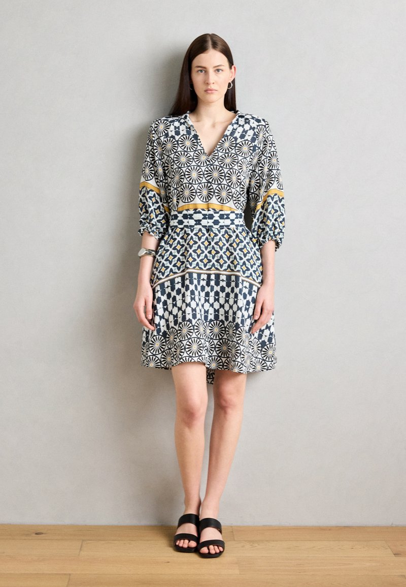 Bedrucktes Kleid mit V-Ausschnitt, Puffärmeln und taillierter Silhouette. Es weist geometrische Muster in Marineblau, Weiß und Gelb auf. Schwarze Sandalen.
