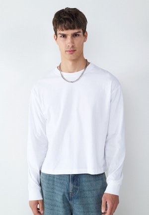 Ung mand med kort brunt hår iført en hvid langærmet skjorte, blå jeans og en sølvkæde mod en ensfarvet hvid baggrund.